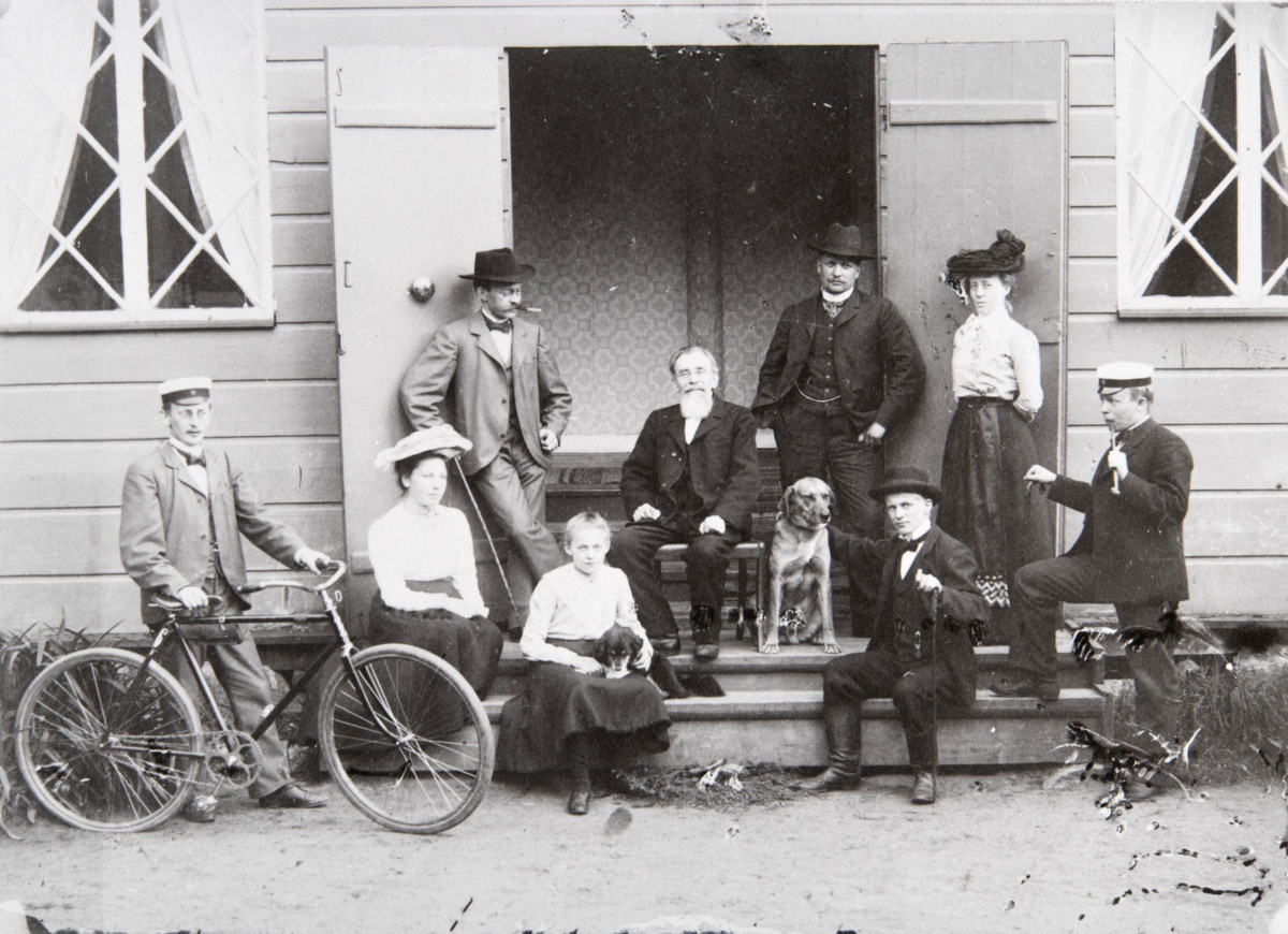 Kirkkoherra A. A. A. Fleege poseeraa kuvassa lastensa Gunnar, Anna, Ragnar, Helge ja Uno Fleegen ja Unon tulevan vaimon Edit Löfbergin kanssa Muuralan pappilan portailla Espoossa vuonna 1903. Kuvaaja: Hans-Peter Sehm / KAMU Espoon kaupunginmuseo 