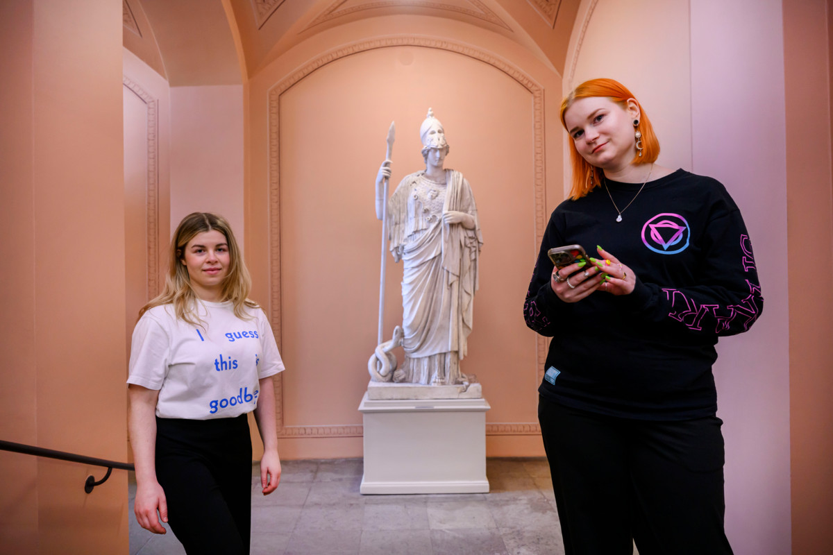 Fanikulttuurista kirjan kirjoittanut Oona Pohjolainen (vas), fanittaja Meri Jaakola ja kreikkalaista Athene-jumalatarta esittävä patsas Helsingin yliopiston päärakennuksessa. Pohjolainen vertaa nykyjulkkiksia antiikin tarustoon: “Kuten antiikin jumalat, myös julkkikset nähdään rikkaina, täydellisinä ja kaikkivoipina, mutta heistä etsitään myös inhimillisyyttä ja haavoittuvaisuutta.” Kuva: Jussi Helttunen.