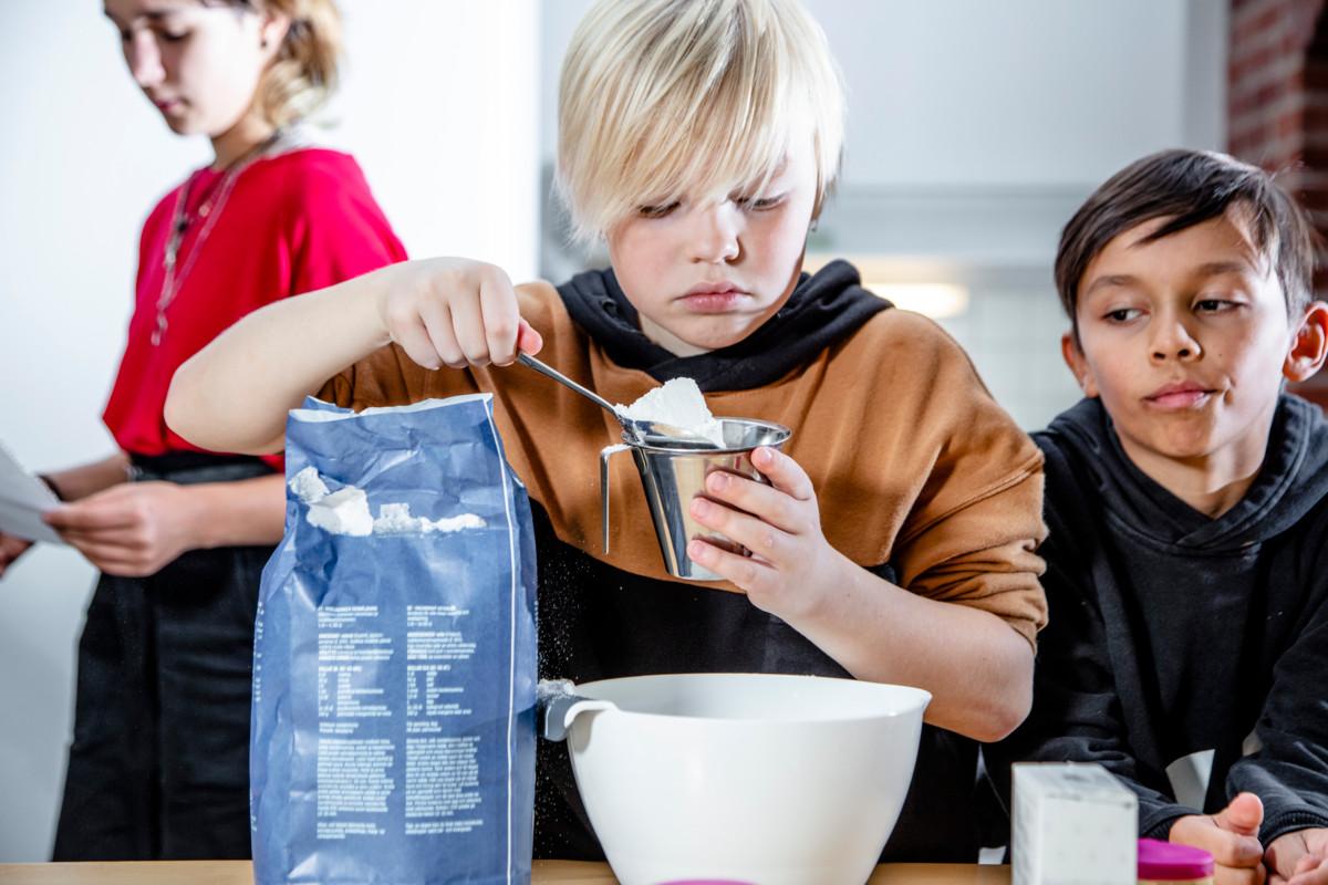 Dani Törmi täyttää vehnäjauhoilla mitta-astiaa. Adi Gupta odottaa omaan vuoroaan. Tara Lankinen lukee reseptistä, mitä seuraavaksi tehdään.