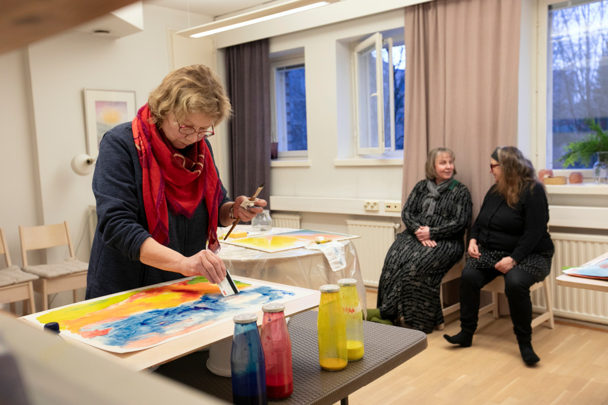 Taina Pulkkinen (vas.) on pannut siveltimen sivuun ja maalaa pankkikortin kokoisella kortilla. Tuija Varjoranta (oik.) juttelee Nina Klemmtin kanssa.