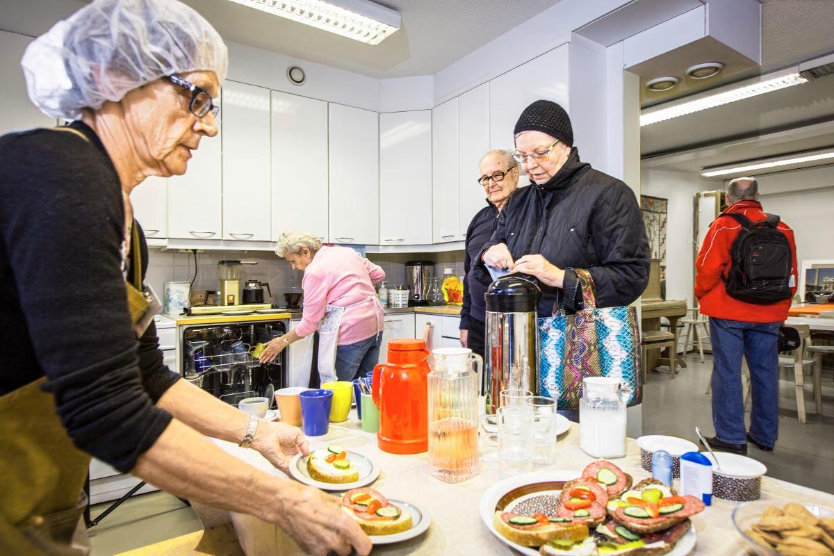 Pukinmäen Porttikahvila toimii vapaaehtoisvoimin: Ulla Antikainen kattaa pöytää ja Raija Välimaa täyttää tiskikonetta. Kahvilan asiakkaat Rauno Hakkarainen ja Tuula Lehto hakevat syötävää ja juotavaa. Kuva: Esko Jämsä