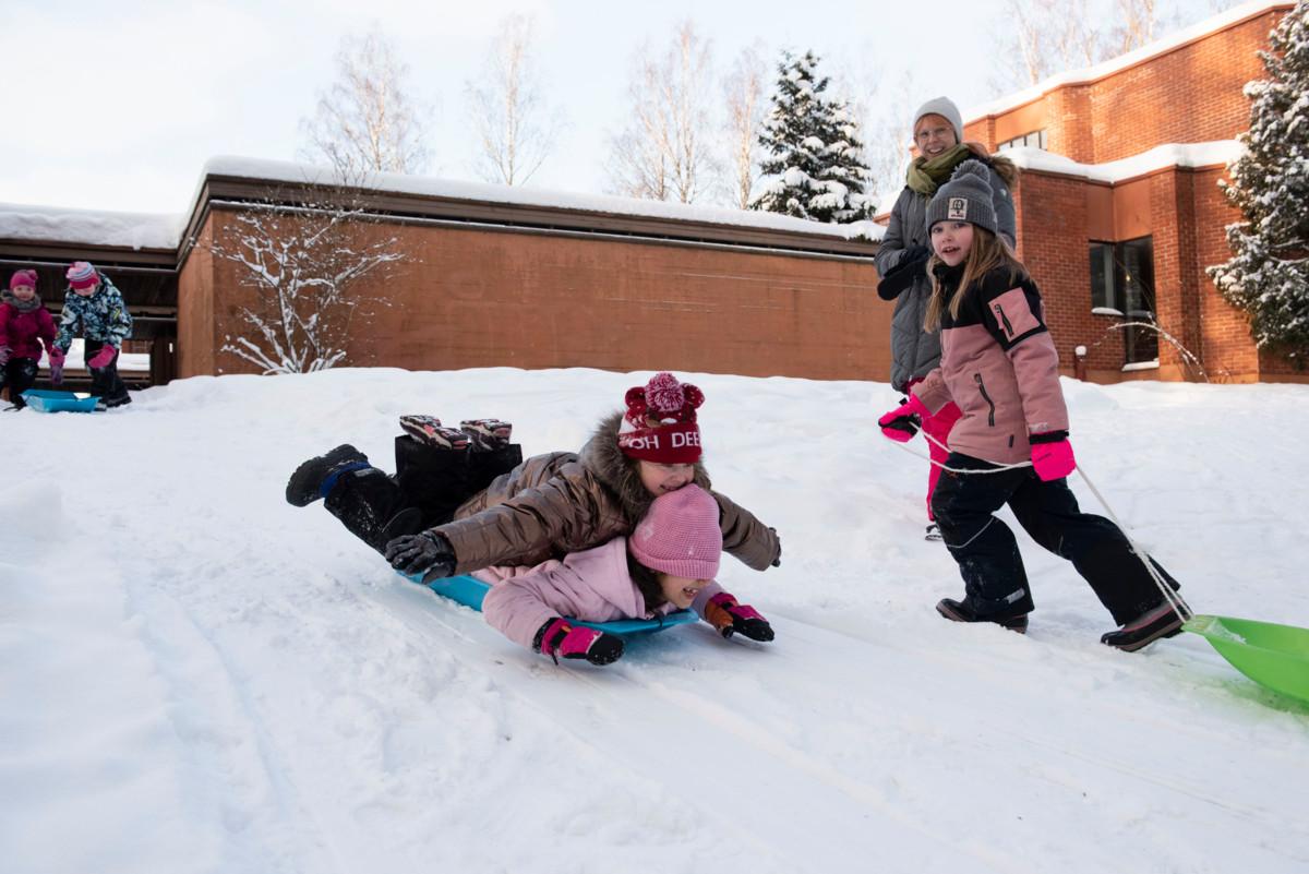 Nathalie Al-zeilaa laskee mäkeä Kivistön kirkon pihalla selässään toinen iltapäiväkerholainen. Vasemmalla Tinka Hänninen ja Emma Skyttä valmistautuvat lähtöön. Mäkeä nousemassa Ilona Siirtola. Lastenohjaaja Satu Karonen seuraa lasten ilakointia.