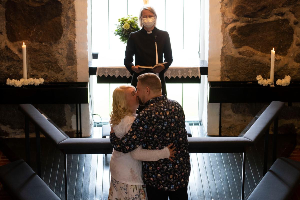 Pappi Kaisa Kariranta hymyilee maskin takana, kun Sari Henriksson ja Tommi Kangas vaihtavat alttarilla suudelman.