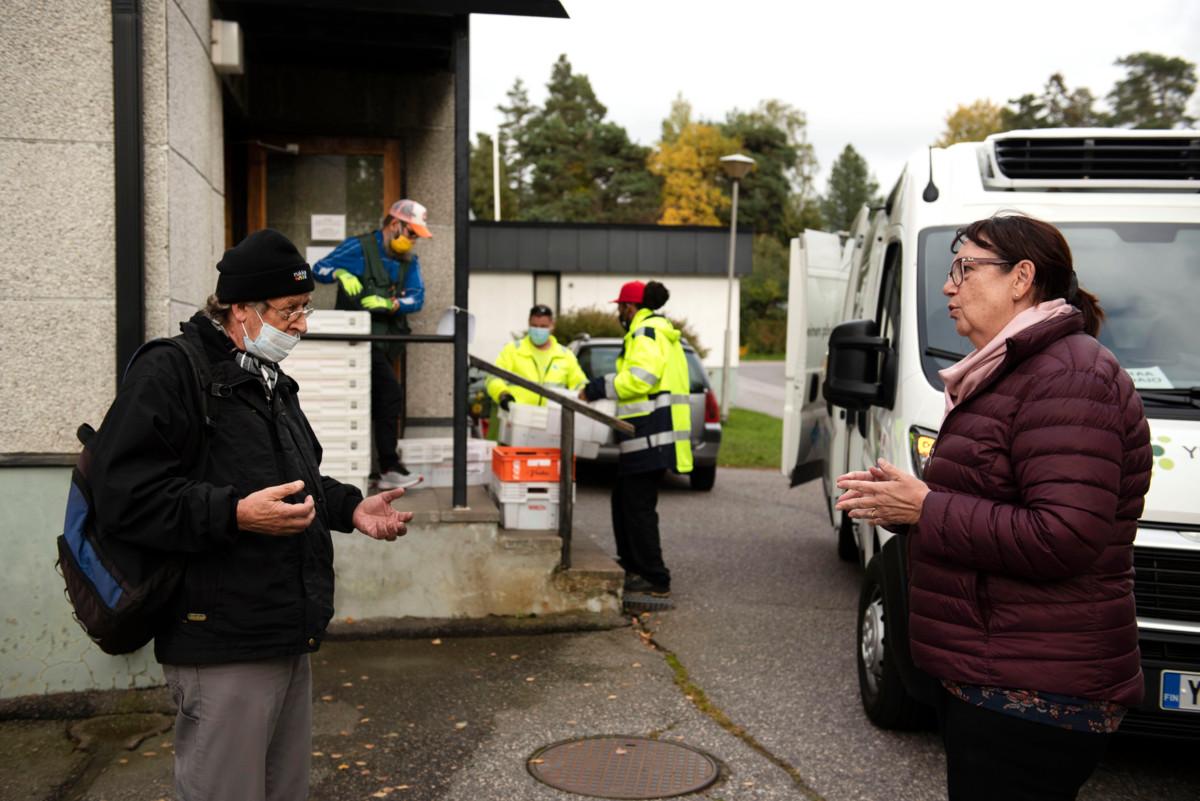 Vahti ry:n puheenjohtaja Tiina Tuomela juttelemassa yhdistyksen ruokajakelusta apua saavan Kaken kanssa. Takana Vantaan yhteisen pöydän auto tuo ruokatoimitusta lajiteltavaksi.